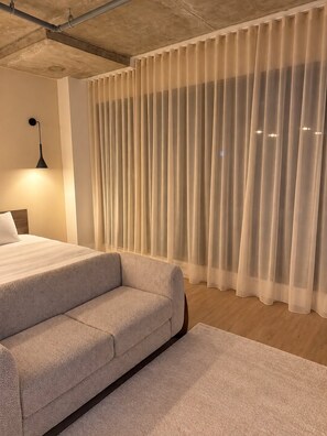 Quarto moderno com cama, sofá, luminária e cortinas leves.