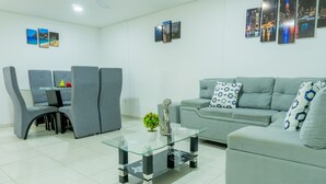 A modern living room with a grey sofa, a glass coffee table, and grey armchairs. There are wall-mounted picture frames and a small table with decorative items.