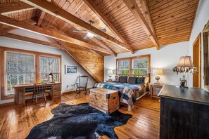 A spacious loft bedroom with a sloped ceiling, wooden beams, large windows, a desk, a bed with a quilt, and a cowhide rug.