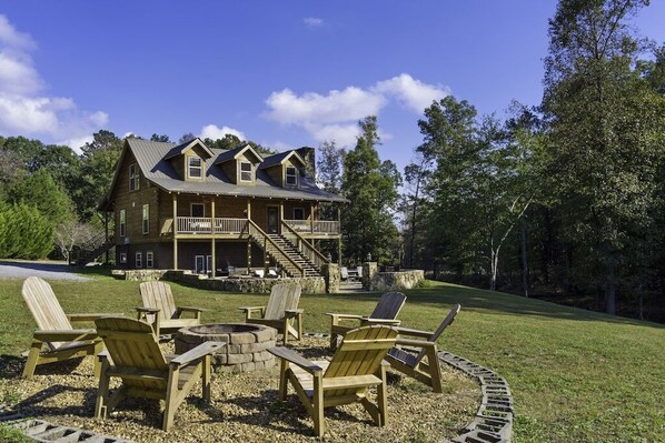 A large wooden house with a balcony and a stone fireplace surrounded by Adirondack chairs.