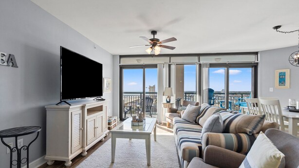 A living room with a flat-screen TV, a ceiling fan, a large window, and a balcony with outdoor furniture. - Condo w/ocean & salt marsh view balcony, steps to the beach w/fitness center, indoor & outdoor pools (North Myrtle Beach)