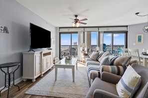 Una sala de estar con televisor de pantalla plana, ventilador de techo, ventana grande y balcón con mobiliario exterior.