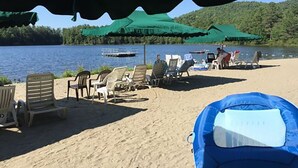 Een strand met meerdere ligbedden en parasols, een boot op het water, en een beboste heuvel op de achtergrond.