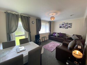 A living room with a dining table, chairs, a sofa, and a TV. There are curtains, a clock, and wall art. - Lands End (Liskeard)