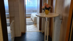 A narrow hallway with a small round table and a vase of flowers. The hallway leads to a room with a bed and a window with curtains. - Apartment  (Basilea)