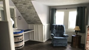 Una habitación con techo inclinado, un sillón azul, un barril de madera, una ventana con cortinas y una lámpara sobre una mesita auxiliar.