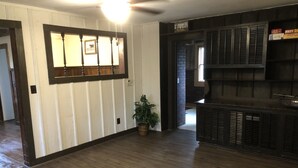 Una habitación amplia con suelo de madera, un ventanal con persianas, una zona de bar integrada y una planta en maceta.