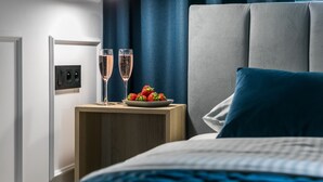 Un letto rifatto con cura, coperto da una coperta blu, un comodino con due bicchieri da champagne e un piatto di fragole, e una moderna testiera.