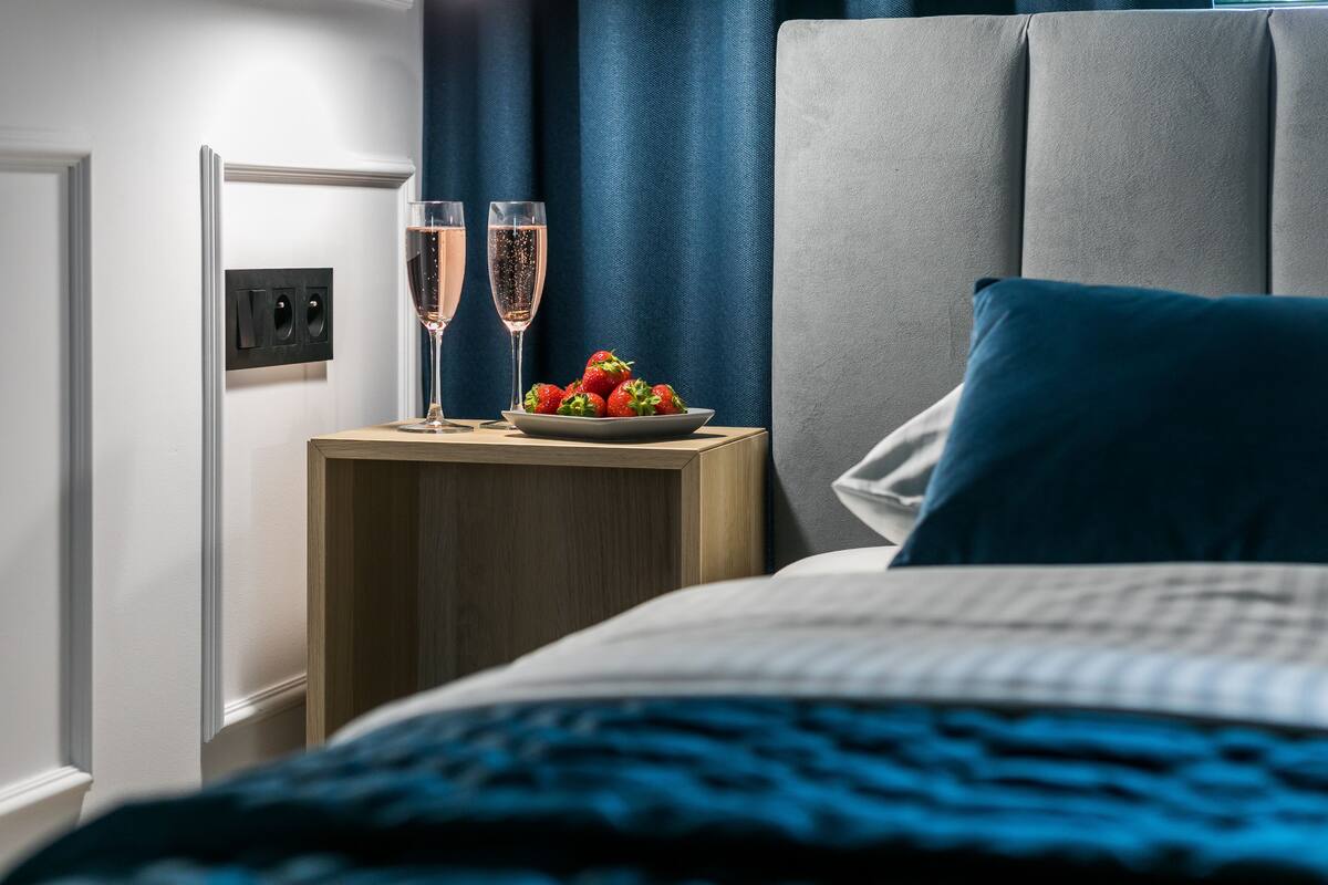 A neatly made bed with a blue blanket, a bedside table with two champagne glasses and a plate of strawberries, and a modern headboard.