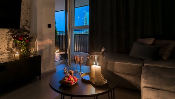 Un soggiorno moderno con un divano in pelle scura, un tavolino tondo con candele, un televisore montato su una parete strutturata e un balcone con vista sullo skyline della città.
