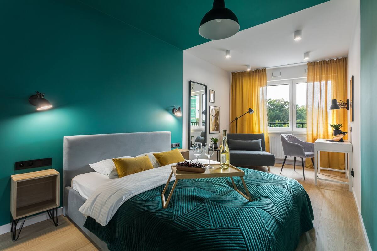 A modern hotel room with teal walls, a large bed with a green blanket, a bedside table with a bottle and glasses, a desk with a chair, and a view of the outdoors through a window with yellow curtains.