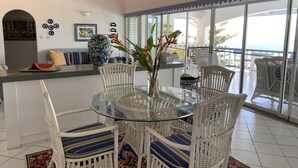 A dining area with a glass table surrounded by white wicker chairs with blue cushions, a large decorative vase with flowers, and a view of the ocean through large windows.