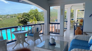 A balcony with wicker furniture, a glass table, and a view of a lush landscape and a pool.
