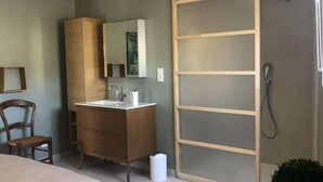 Une chambre moderne comprenant un lit, une commode en bois, une coiffeuse avec un miroir et une porte coulissante en verre donnant sur un espace douche.