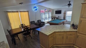 A room with a pool table, a dining table with chairs, a kitchen area with wooden cabinets, and a ceiling fan. - Parker, AZ - Hilltop Hideaway (Parker)