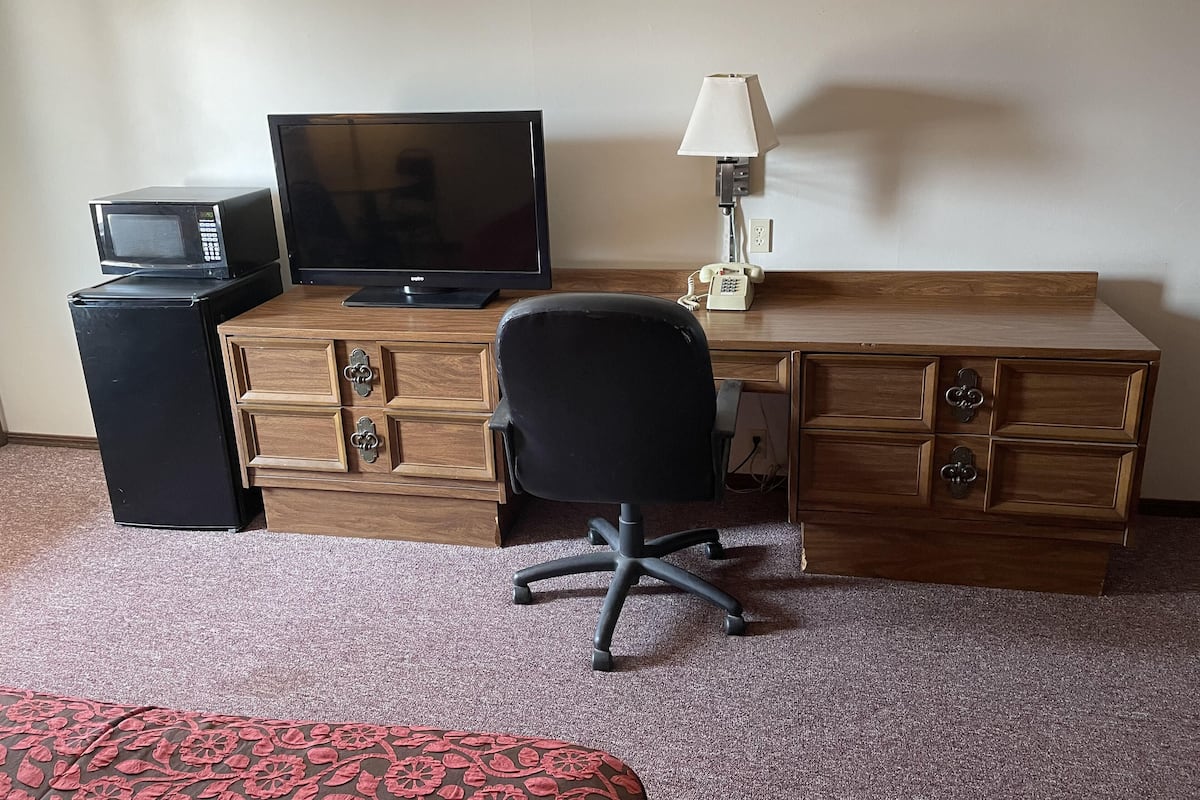 A hotel room with a wooden desk, a black office chair, a television, a microwave, and a telephone.