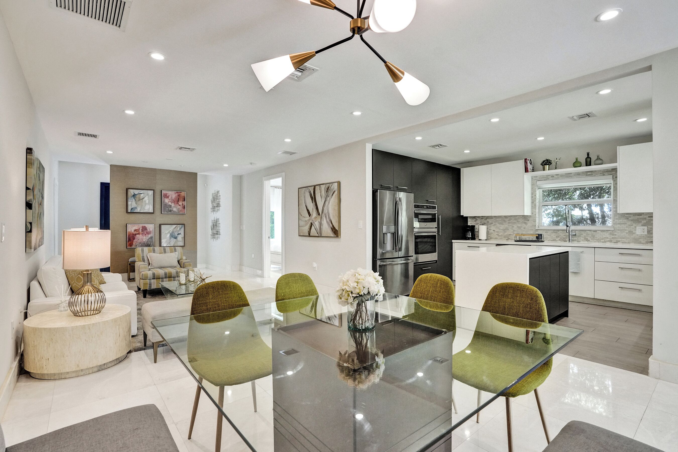 Eine moderne Küche mit Edelstahlgeräten, ein Essbereich mit Glastisch und grünen Stühlen sowie ein Wohnbereich mit Sofa und Lampe.