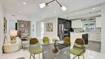 Eine moderne Küche mit Edelstahlgeräten, ein Essbereich mit Glastisch und grünen Stühlen sowie ein Wohnbereich mit Sofa und Lampe.