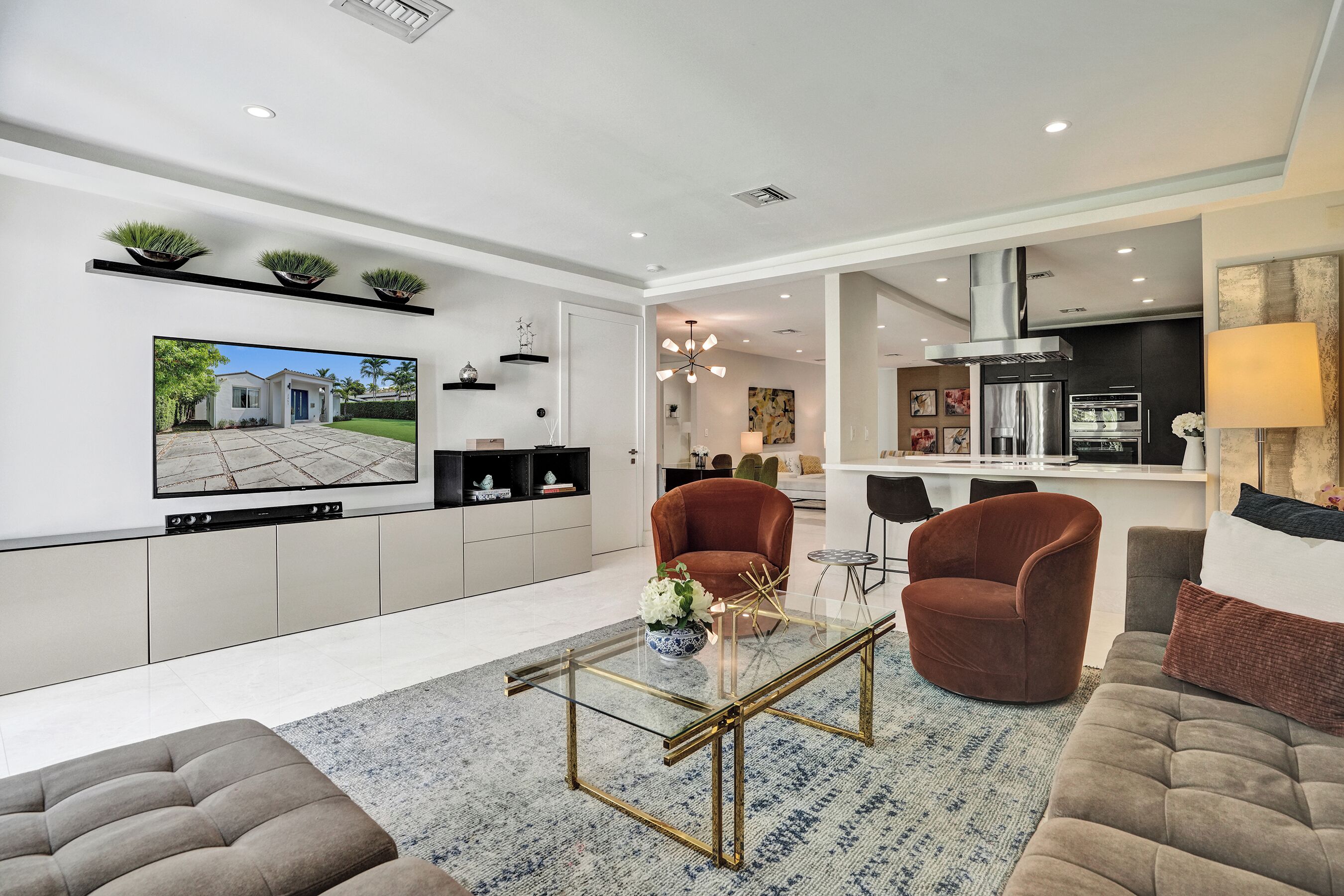 A modern living room with a glass coffee table, two brown armchairs, a sofa, a TV mounted on the wall, and a kitchen in the background.