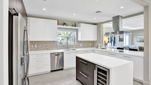 A modern kitchen with white cabinetry, a stainless steel dishwasher, and a large island with a wine cooler. - Renovated 4 Bedroom home, tastefully designed! (Miami)