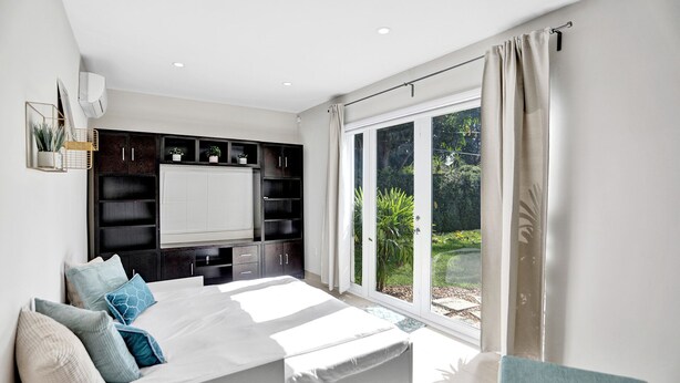 A bedroom with a large bed, built-in shelving, a window with curtains, and a view of greenery outside. - Renovated 4 Bedroom home, tastefully designed! (Miami)