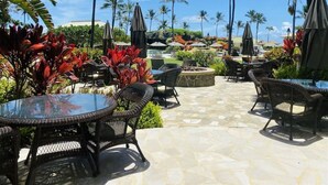 An outdoor patio area with round tables and chairs, surrounded by tropical plants and palm trees. - Breathtaking Oceanfront Resort, 4 pools, 2 Jacuzzis and Restaurants on Site  (Lihue)