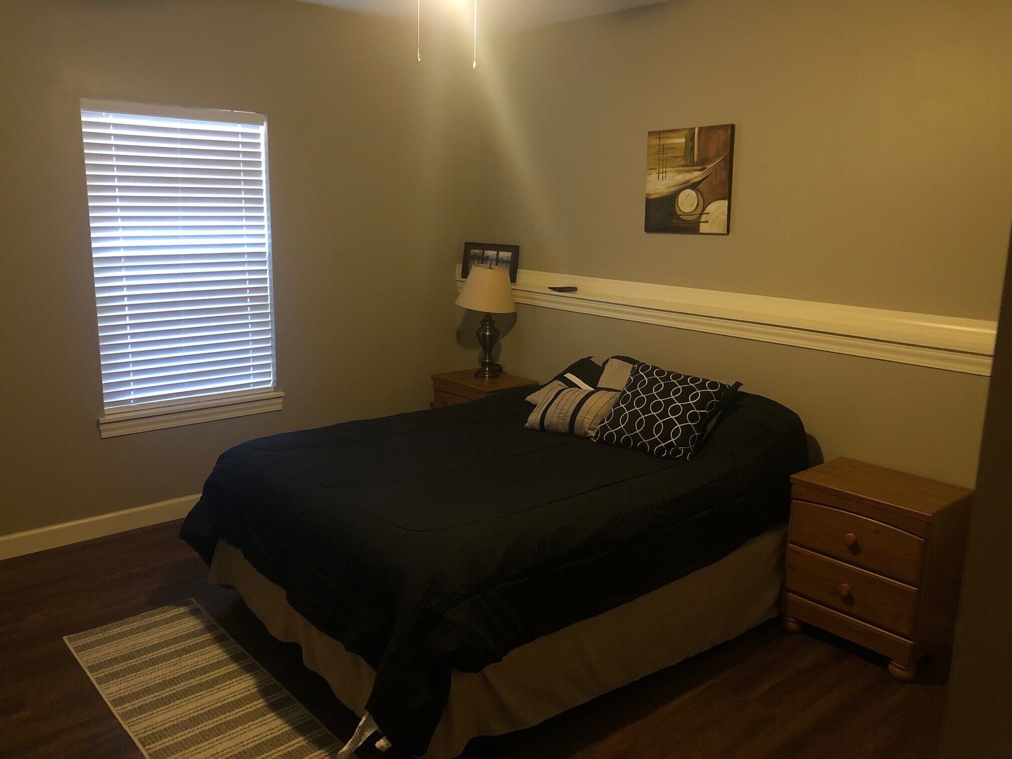 Un dormitorio con cama, mesitas de noche, lámpara, un cuadro y una ventana con persianas.