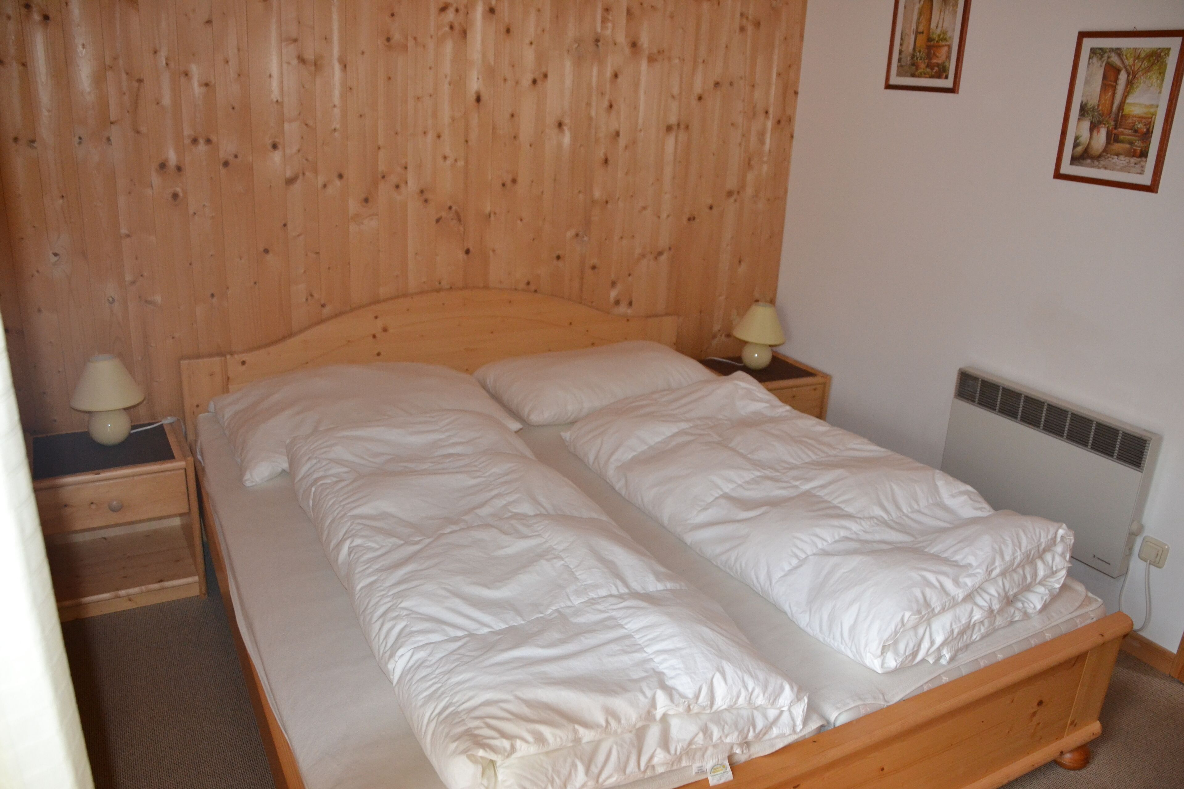 Uma cama de madeira com roupa de cama branca, uma mesa de cabeceira com abajur e um radiador na parede.