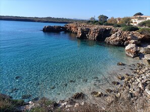 A coastal landscape with clear blue waters, rocky shorelines, and a building with a tiled roof.