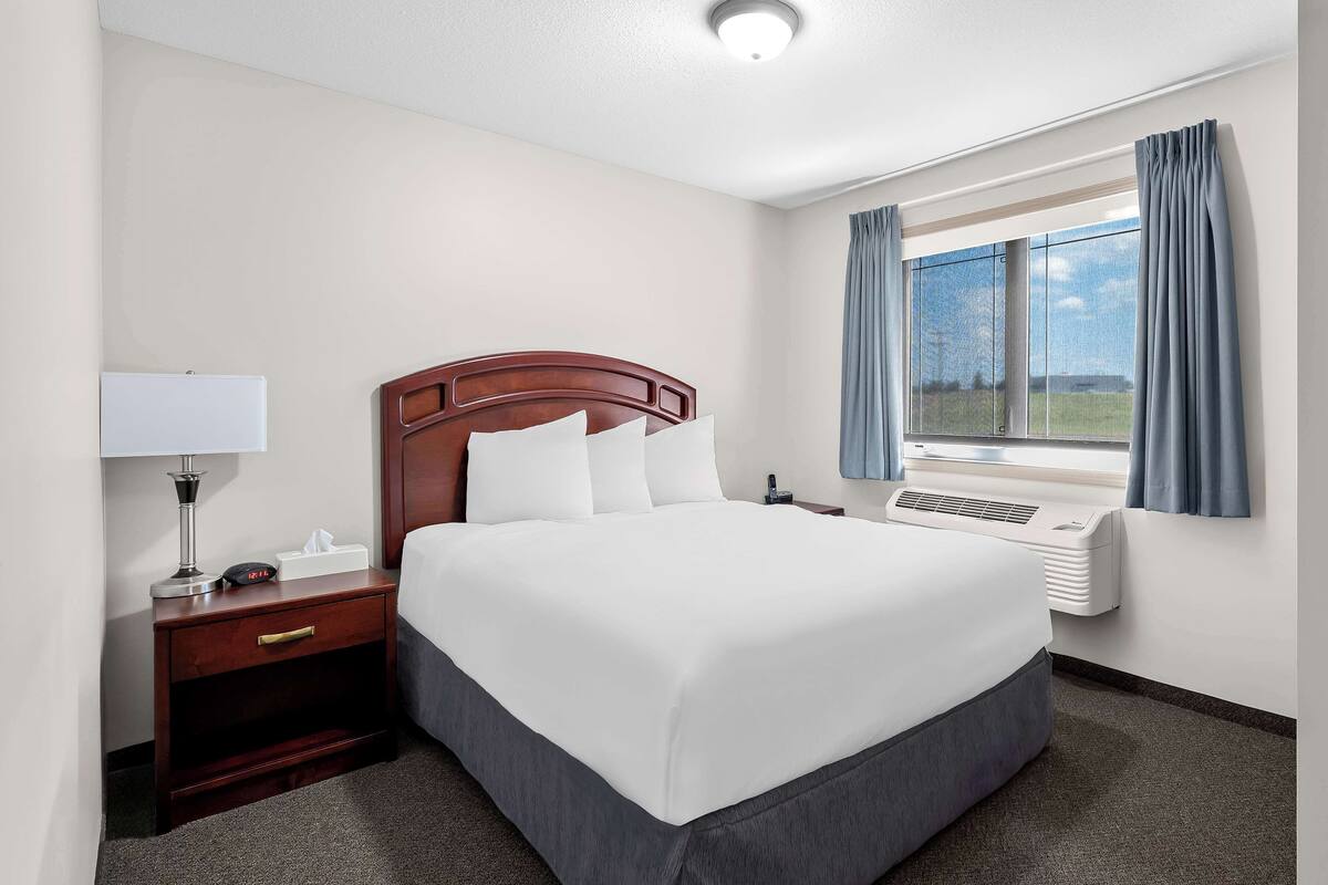 A hotel room with a large bed, a nightstand with a lamp, a window with curtains, and an air conditioning unit.