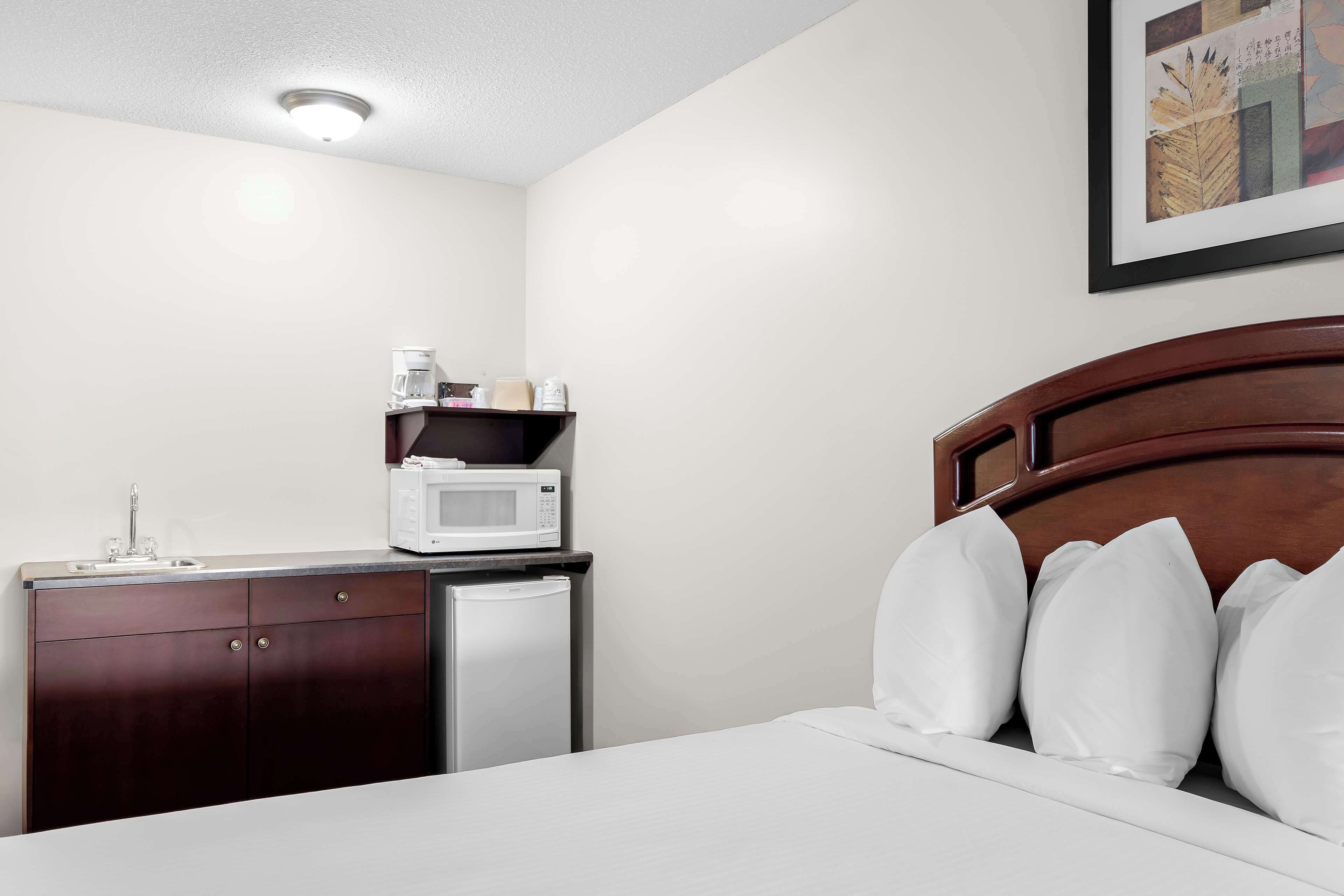 A hotel room with a bed, white pillows, a wooden headboard, a small kitchenette with a microwave, and a refrigerator.