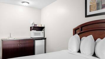 A hotel room with a bed, white pillows, a wooden headboard, a small kitchenette with a microwave, and a refrigerator.