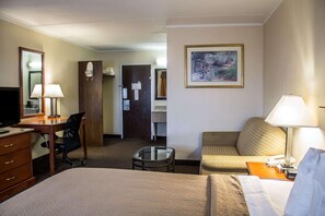 A hotel room with a bed, a desk with a chair, a small table, a sofa, a TV, and a painting on the wall.