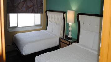 Quarto de hotel com duas camas, janela com persianas, abajur e parede com padrão geométrico.
