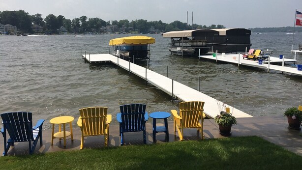 Un muelle a la orilla del lago con un bote cubierto, una fila de sillas Adirondack y una bandera.