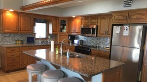 Cocina con gabinetes de madera, encimeras de granito y electrodomésticos de acero inoxidable, y tres taburetes de bar.