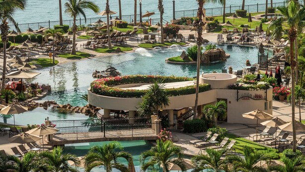 A resort with a large pool area, palm trees, and a view of the ocean.