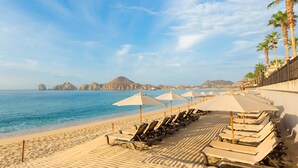 A beach with rows of sun loungers and umbrellas, palm trees, and a clear view of the sea and distant mountains.