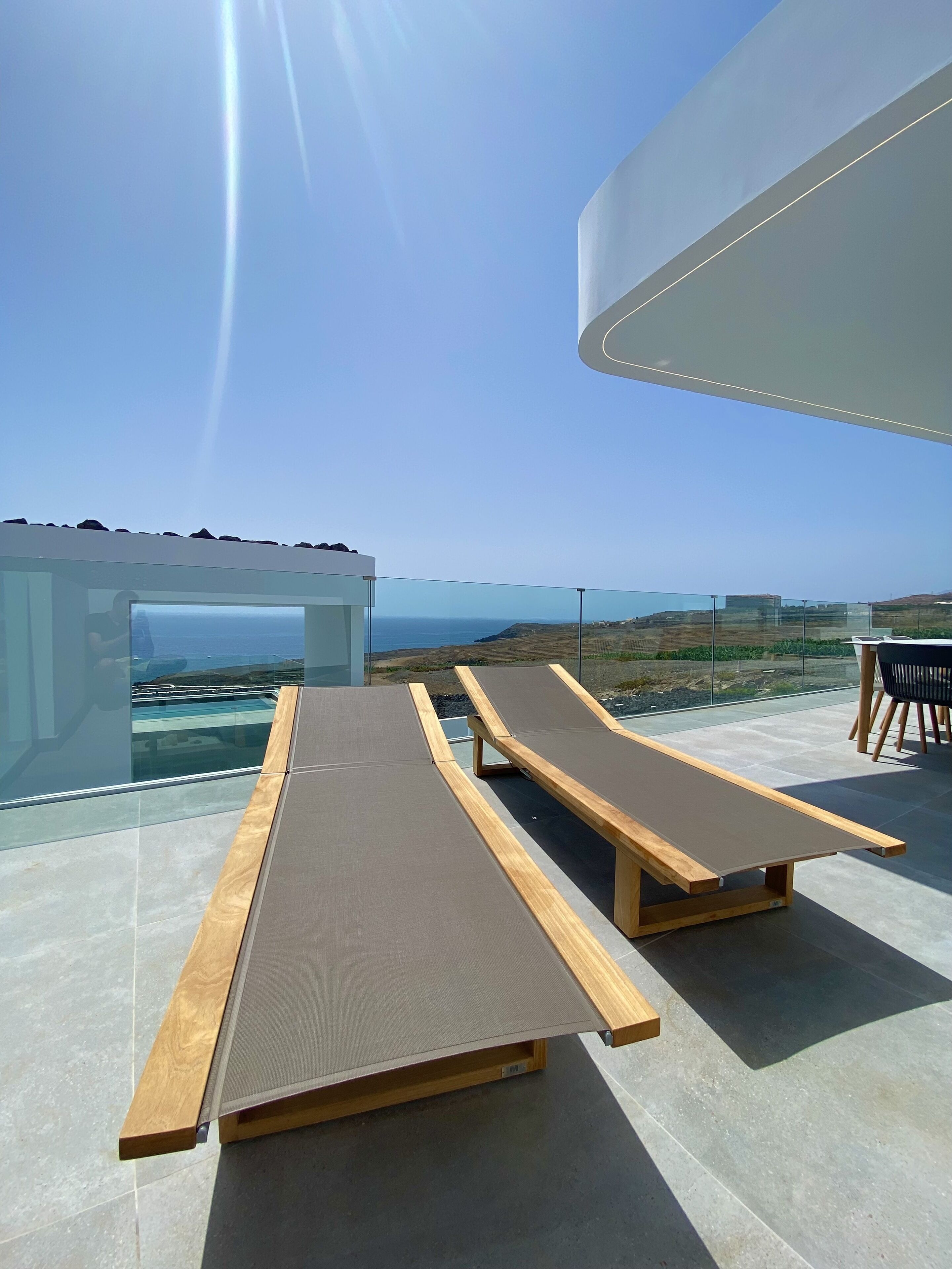 A rooftop terrace with two wooden loungers, a glass balustrade, and a clear view of the ocean and hills.