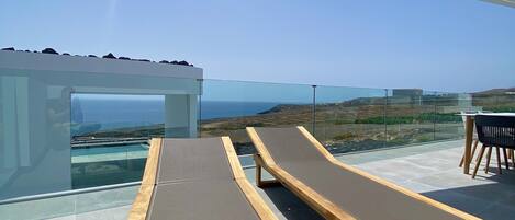 A rooftop terrace with two wooden loungers, a glass balustrade, and a clear view of the ocean and hills.