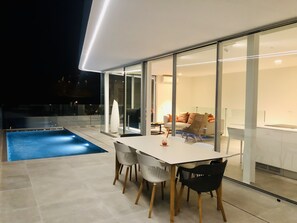 A modern outdoor dining area with a pool, glass walls, and a cozy interior visible through the glass.