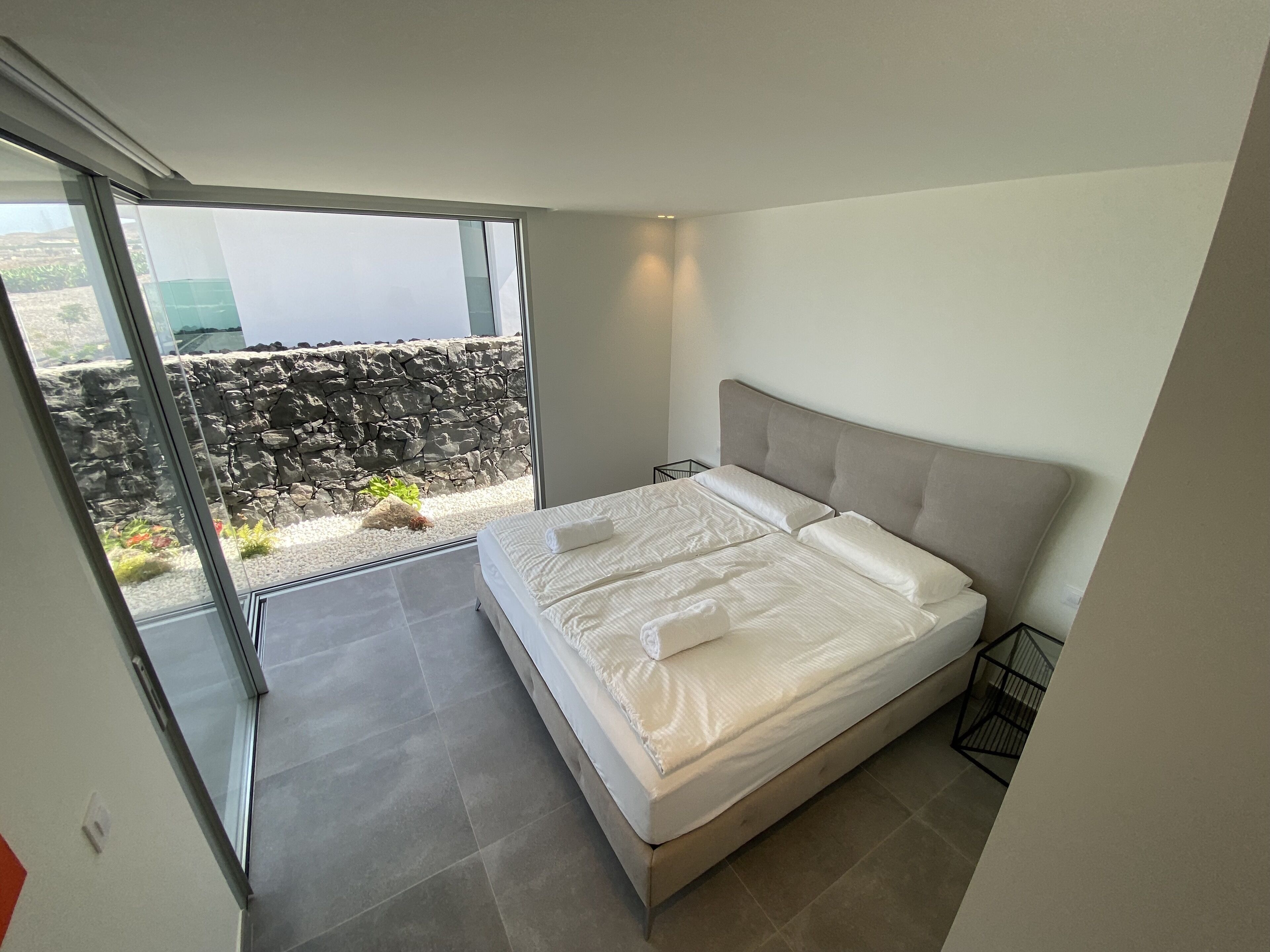 Ein modernes Schlafzimmer mit einem großen Bett, Blick auf eine felsige Landschaft durch eine Glastür und einem Balkon mit einer Steinmauer.