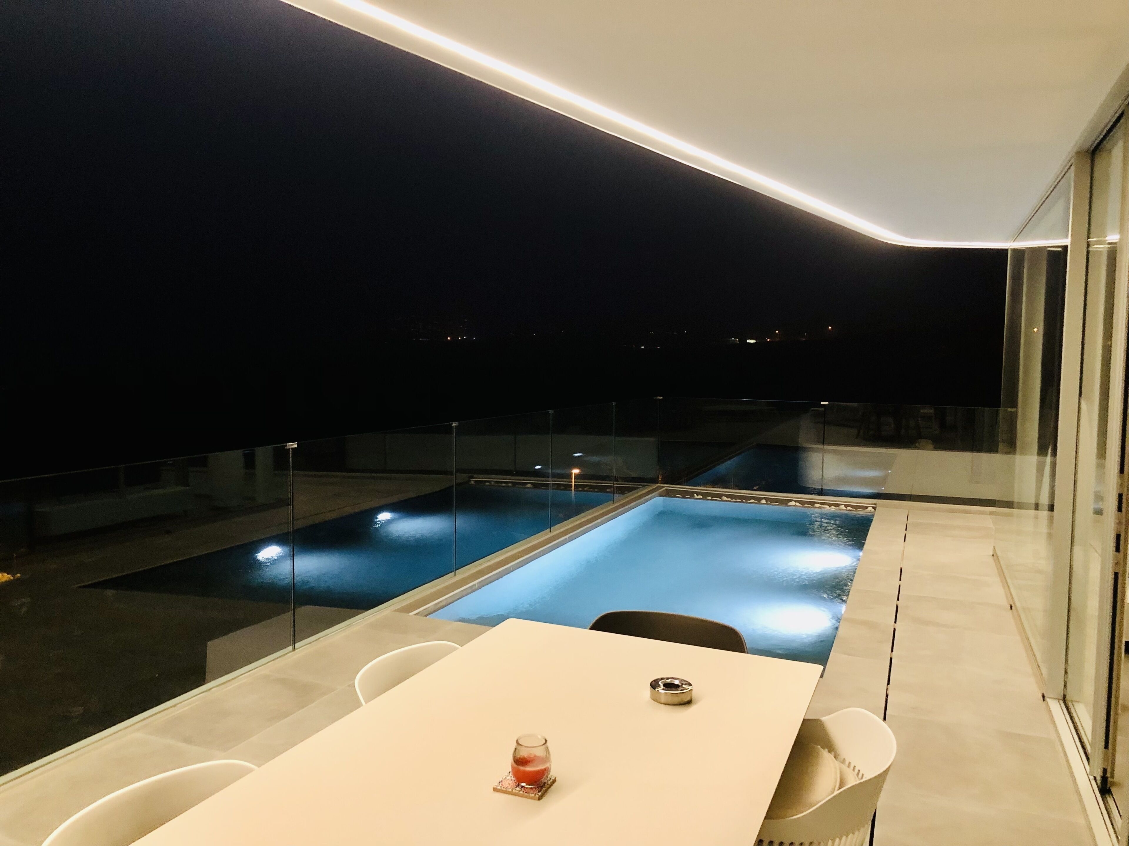 Un espace piscine extérieur moderne, doté d’une balustrade en verre, d’une table avec des chaises et d’un verre contenant une boisson.