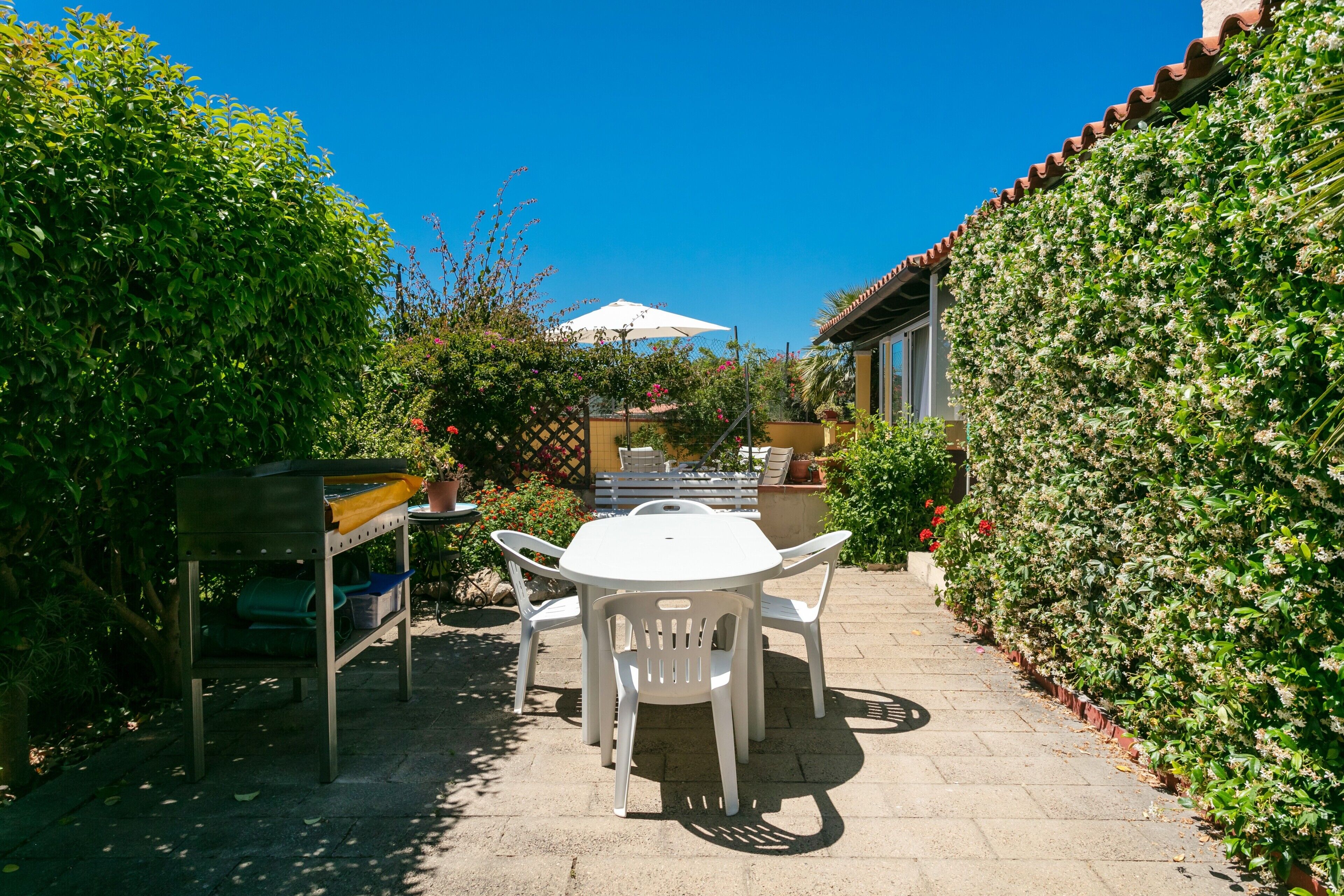 Eine Terrasse mit Tisch und Stühlen, umgeben von Pflanzen und einem weißen Sonnenschirm.