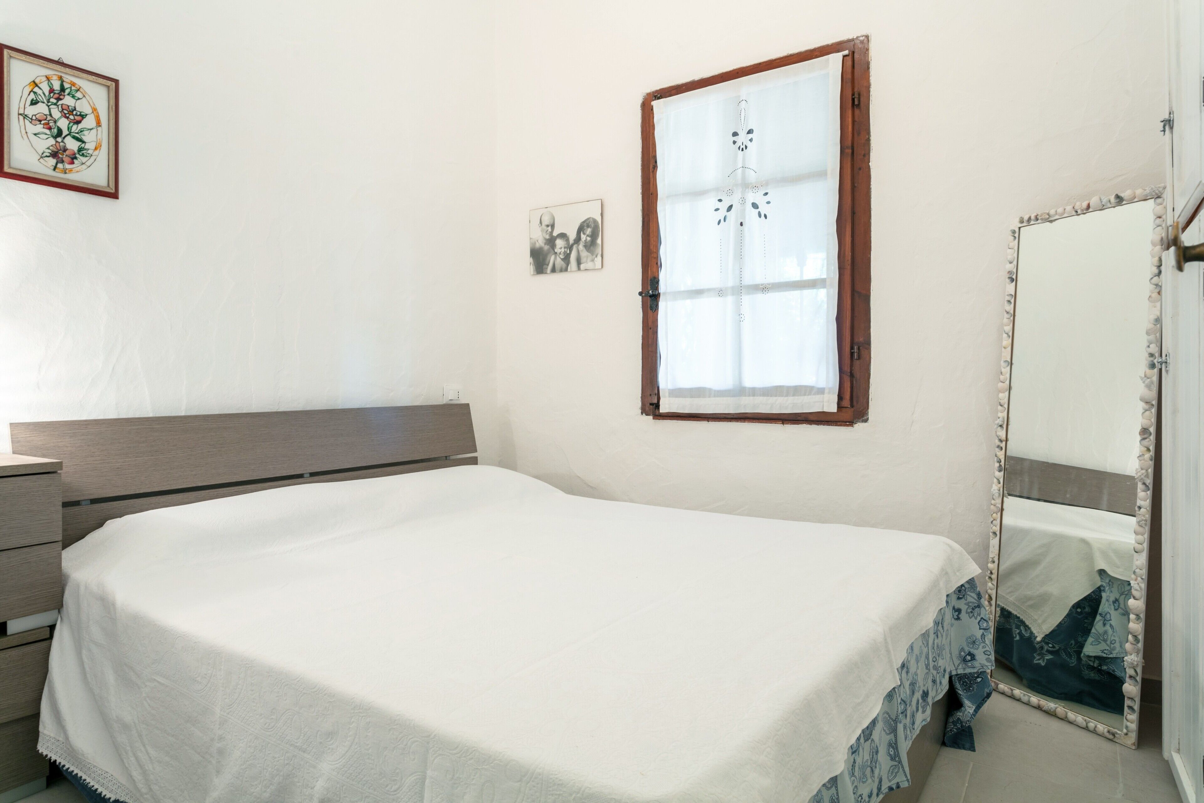 Ein Schlafzimmer mit Bett, Kommode, einem Fenster mit Vorhängen, einem Spiegel und einem Wandbild.