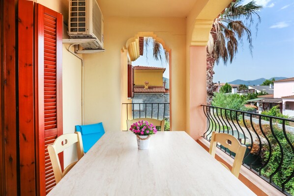 A balcony with a table, chairs, a potted plant, and a view of a building with a tiled roof.