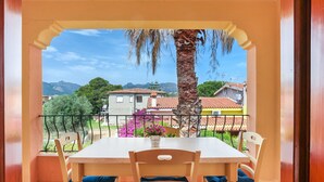 A balcony with a table and chairs, a view of houses and mountains, and a palm tree. - Holiday Apartment "Bilocale Geranium Beach" with Terrace & Air Conditioning (San teodoro)