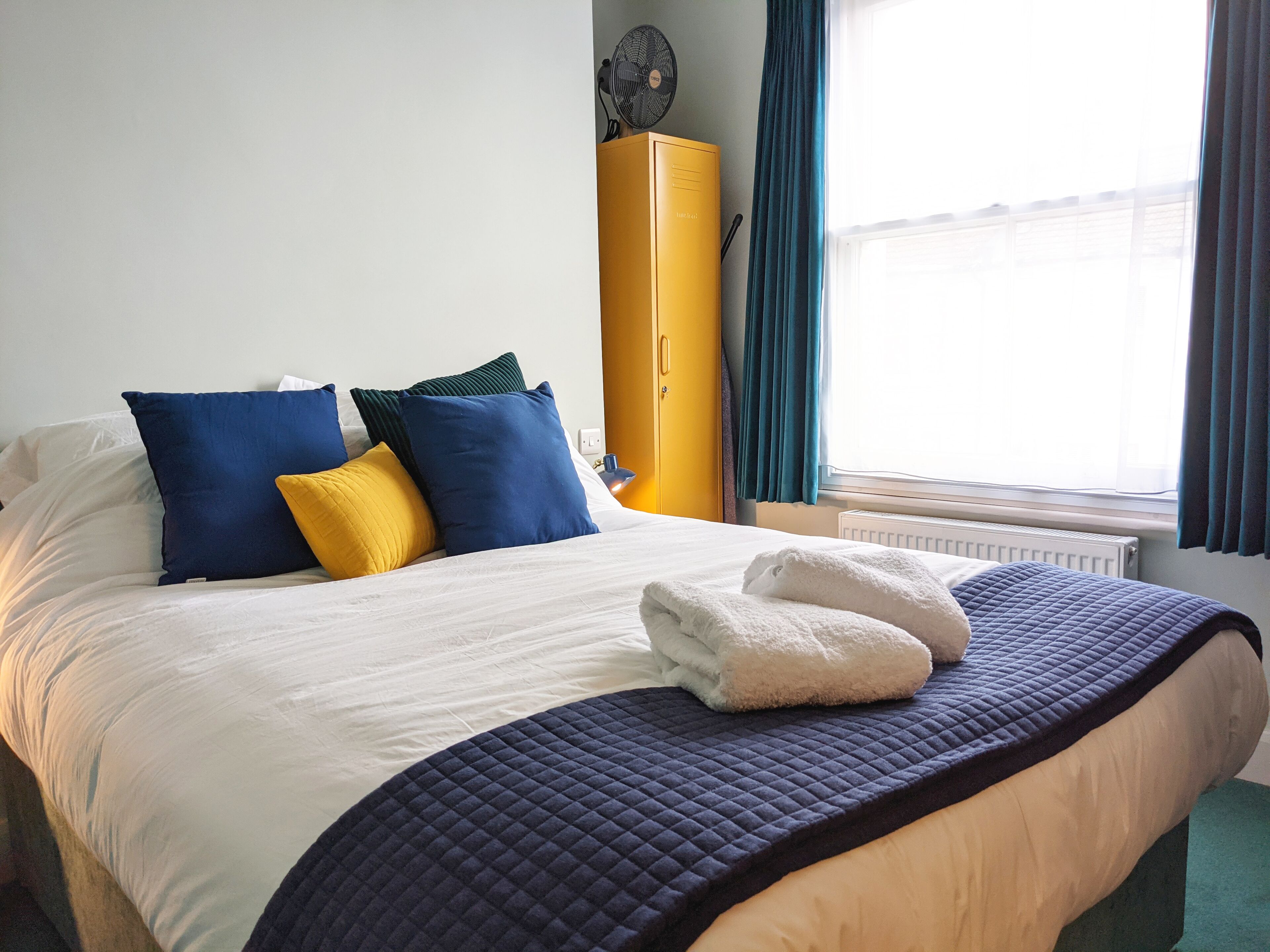 A neatly made bed with a quilted cover, multiple pillows in blue and yellow, and a pair of folded towels on the bed.