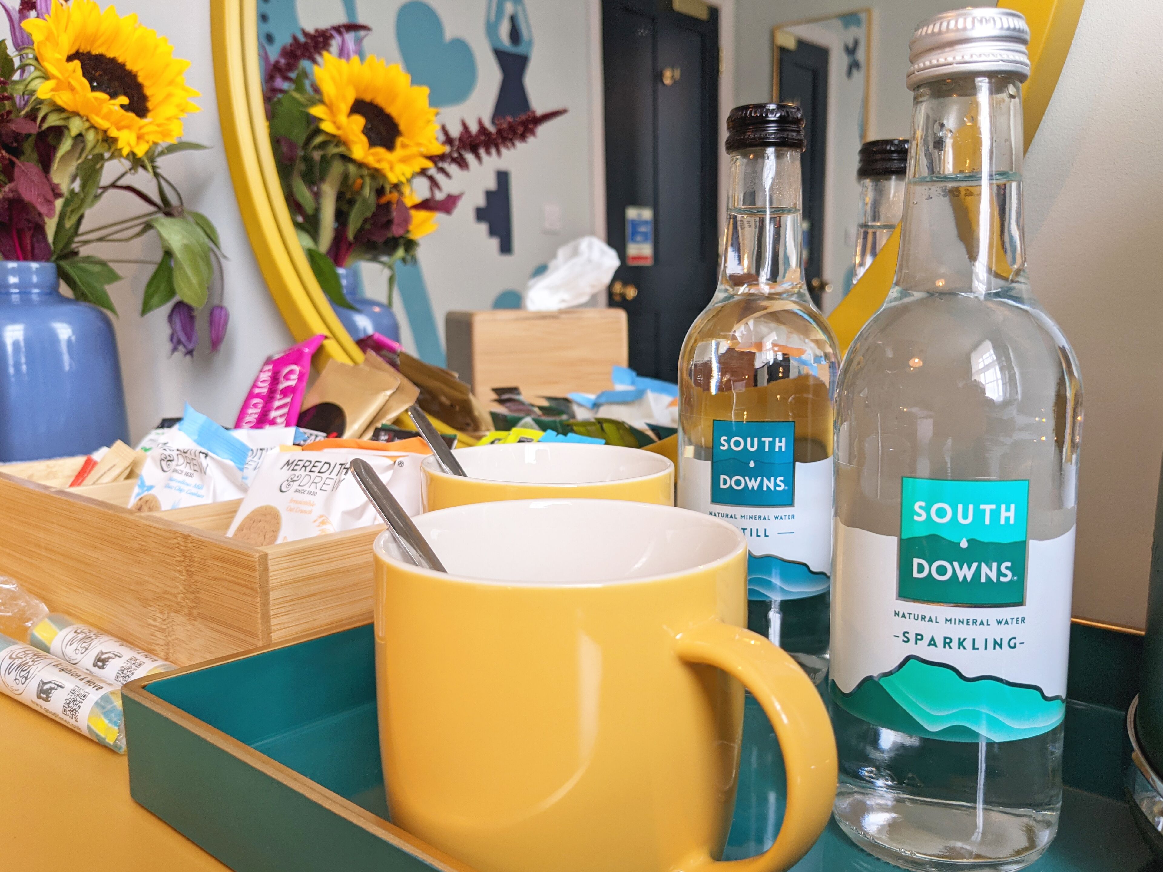 Ein Tisch mit einer gelben Tasse, zwei Flaschen Sprudelmineralwasser von South Downs, einer Holzkiste und einer Vase mit Sonnenblumen.