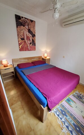 A bedroom with a wooden bed, a nightstand with a lamp, a ceiling fan, and a wall-mounted air conditioner.